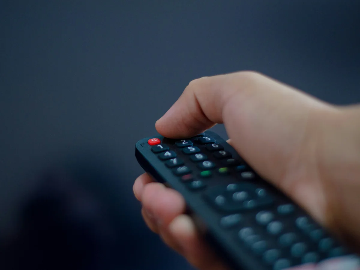 A hand using a television remote against a blue backdrop, illustrating practical viewing guides